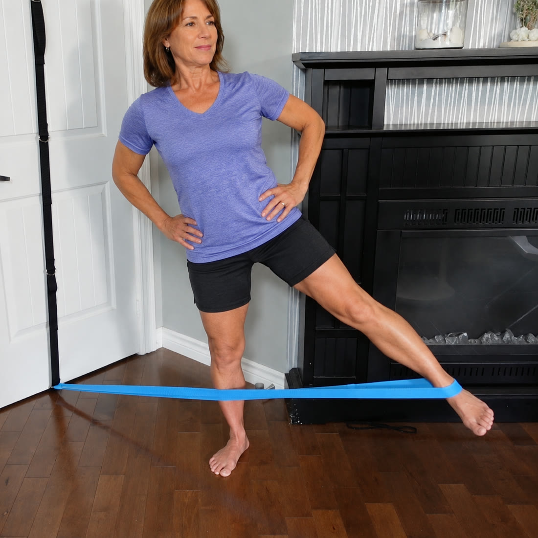 A woman standing with a blue band around her ankle, lifting one leg out to the side